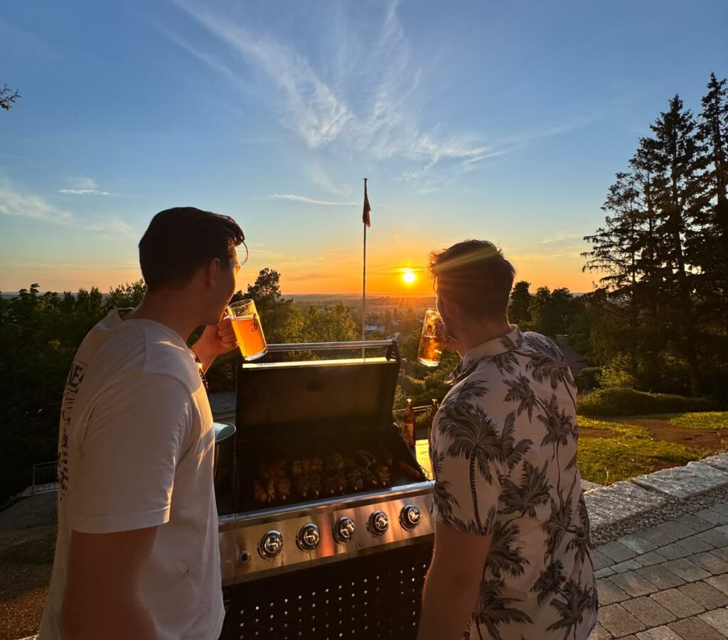 Zwei Männer stehen am Grill und bestaunen bei einem Bier den Sonnenuntergang